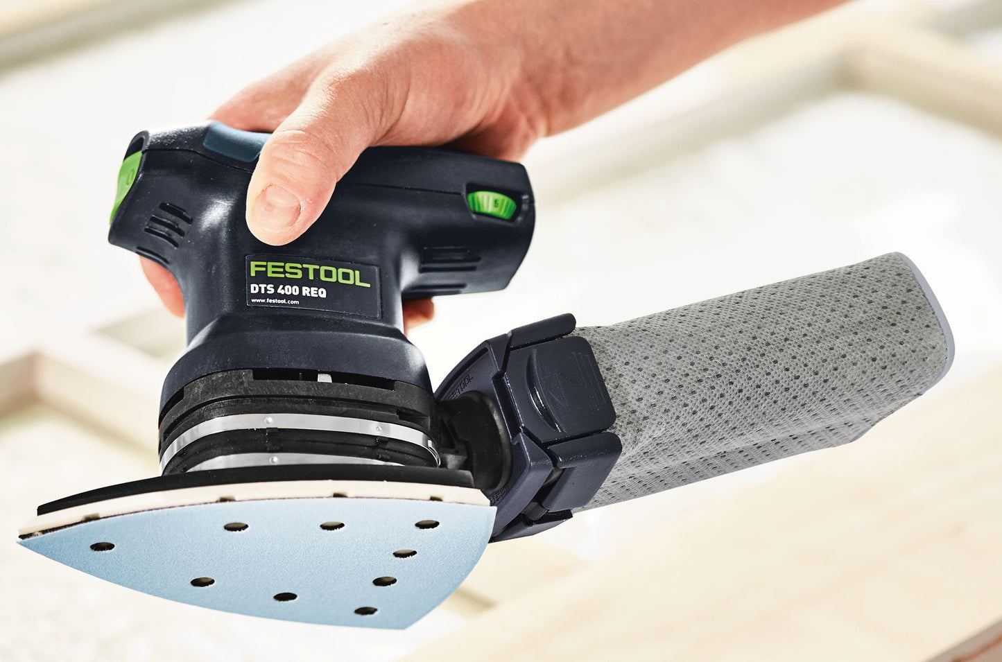 Hand holding Festool electric finishing sander with attached dust collection bag over wooden surface in woodworking tools setting