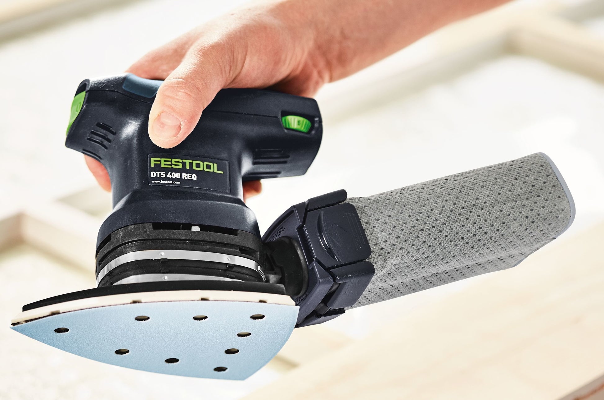 Hand holding Festool electric finishing sander with attached dust collection bag over wooden surface in woodworking tools setting