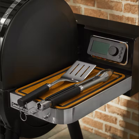 Stainless steel grill spatula and tongs with black soft grip handles resting on a smoker side tray in outdoor living collection