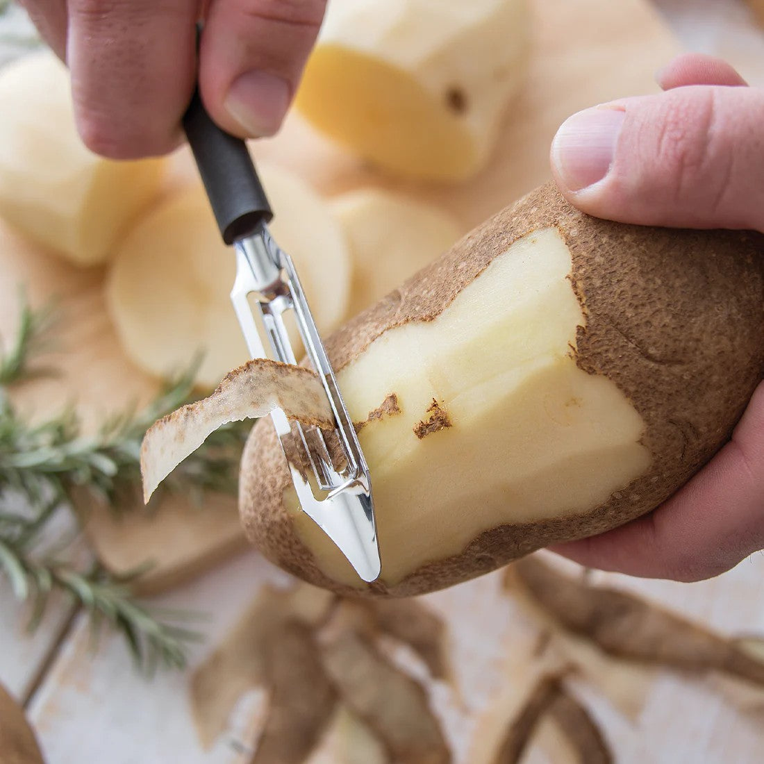 Hand using black-handled peeler to remove skin from potato with peeled potatoes and rosemary in kitchenware setting
