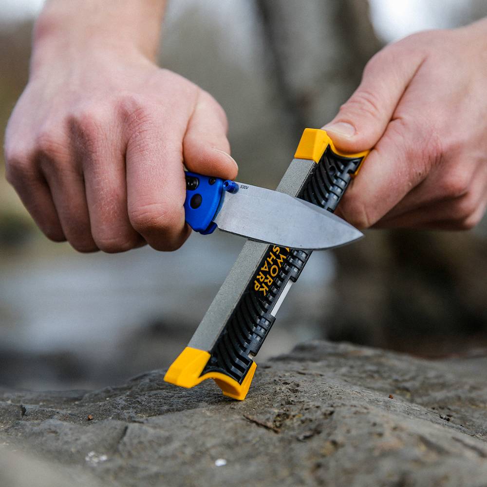 Person sharpening a blue-handled folding knife with a compact black and yellow handheld sharpener on a stone surface