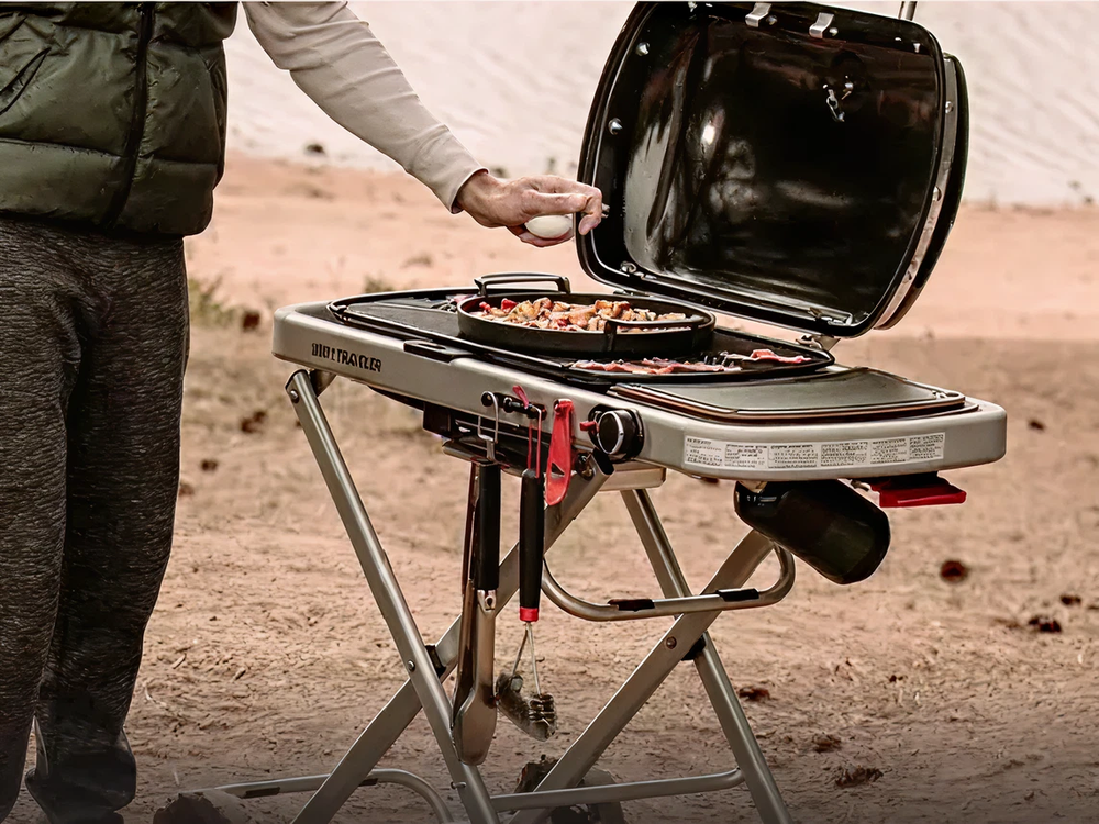 Person seasoning food cooking on a portable gas grill with a lid open, set up outdoors near a sandy or dirt surface.