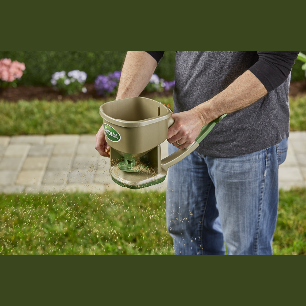 Hand-powered spreader with green handle and brown container distributing lawn seed outdoors in garden tools collection