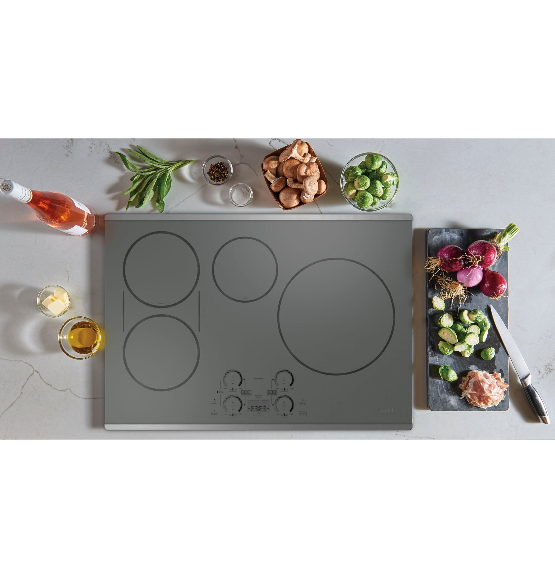 Top view of a sleek built-in induction cooktop with touch controls surrounded by fresh herbs, vegetables, and cooking ingredients