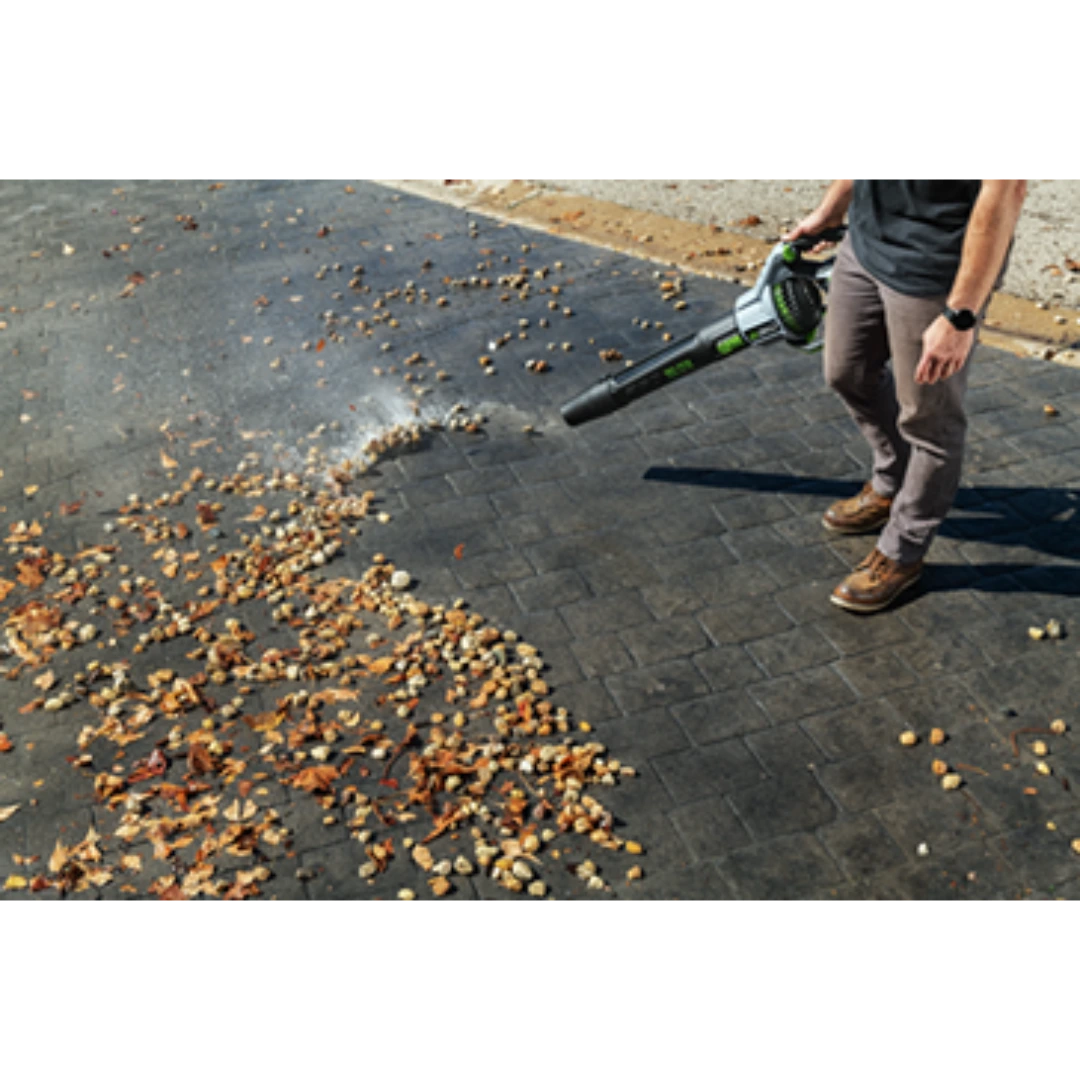 Person using EGO Power+ battery powered blower clearing dry leaves on paved surface in outdoor power tools collection