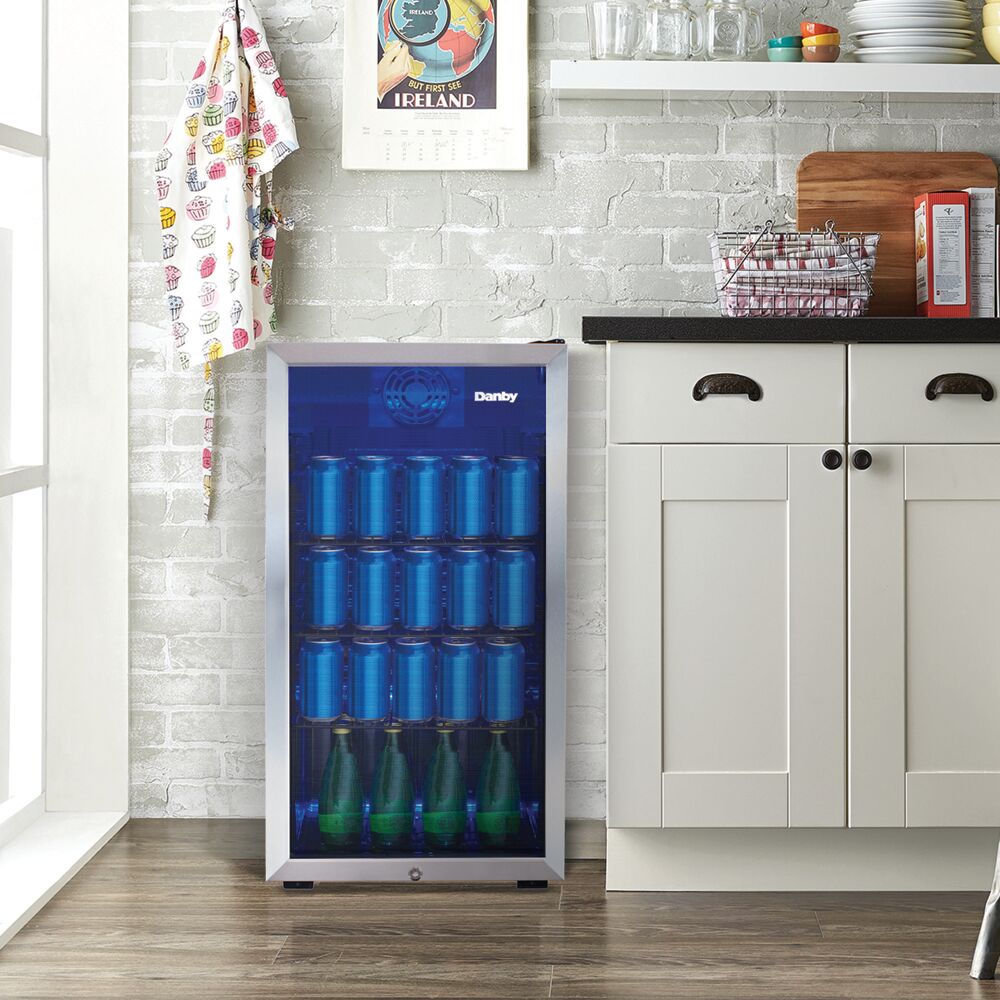 Freestanding beverage center with tinted glass door, wire shelves stocked with blue cans and green bottles in modern kitchen setting