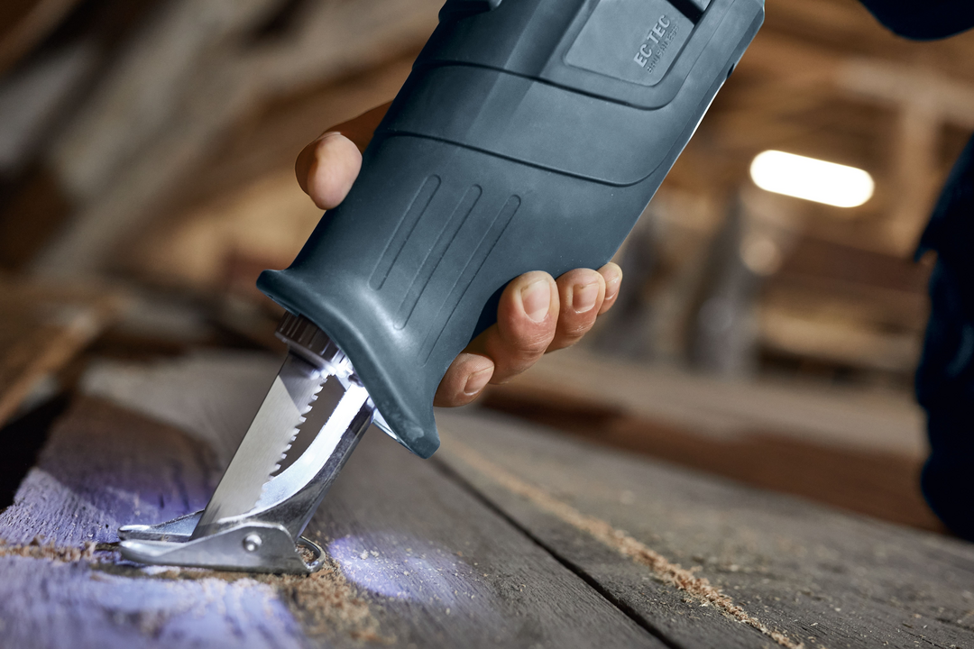 Close-up of hand holding cordless reciprocating saw cutting metal hinge on wooden surface in workshop environment