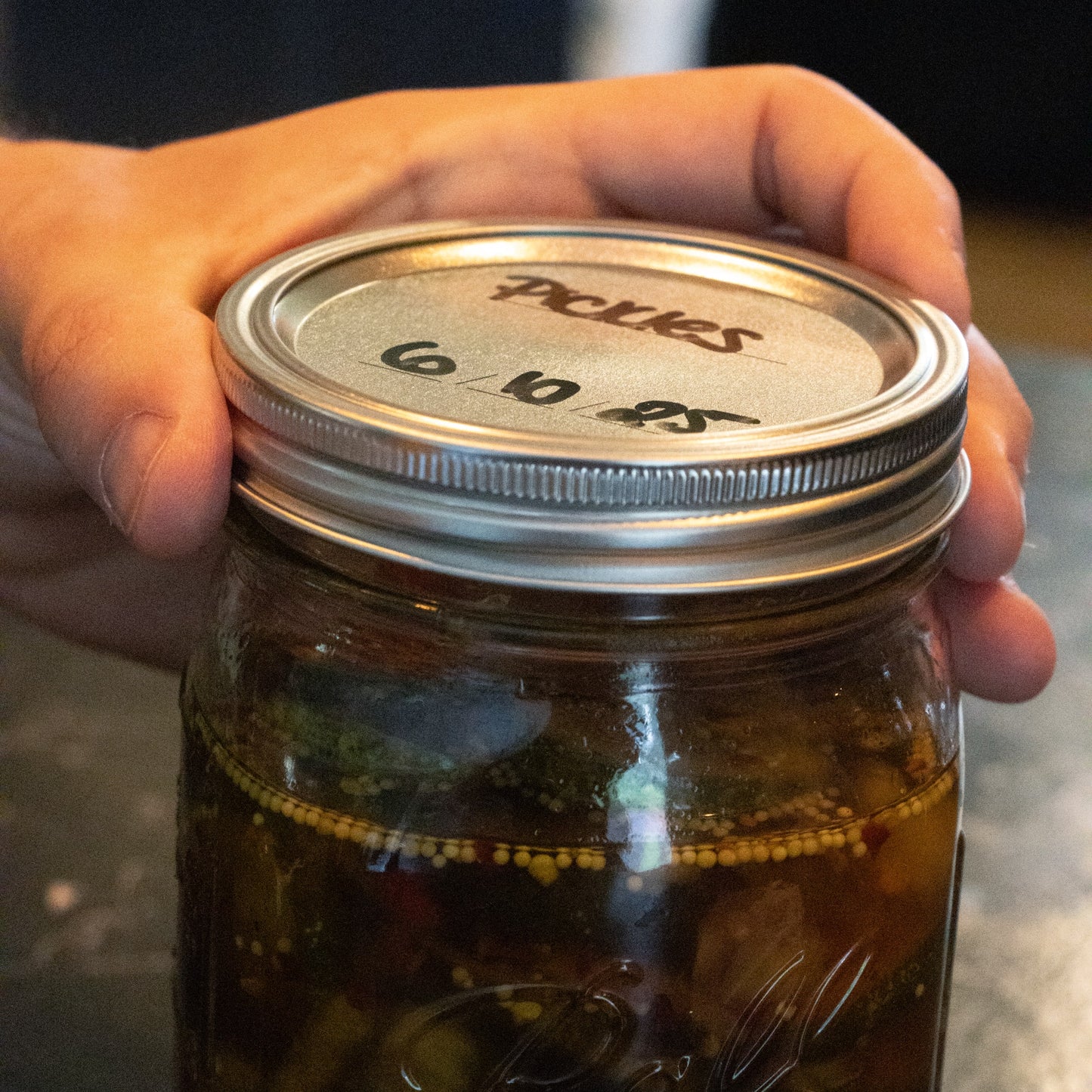 Close-up of a hand holding a glass wide-mouth jar sealed with a silver metal lid labeled with handwritten contents and date, suitable for Kitchenware and Under $50 collections