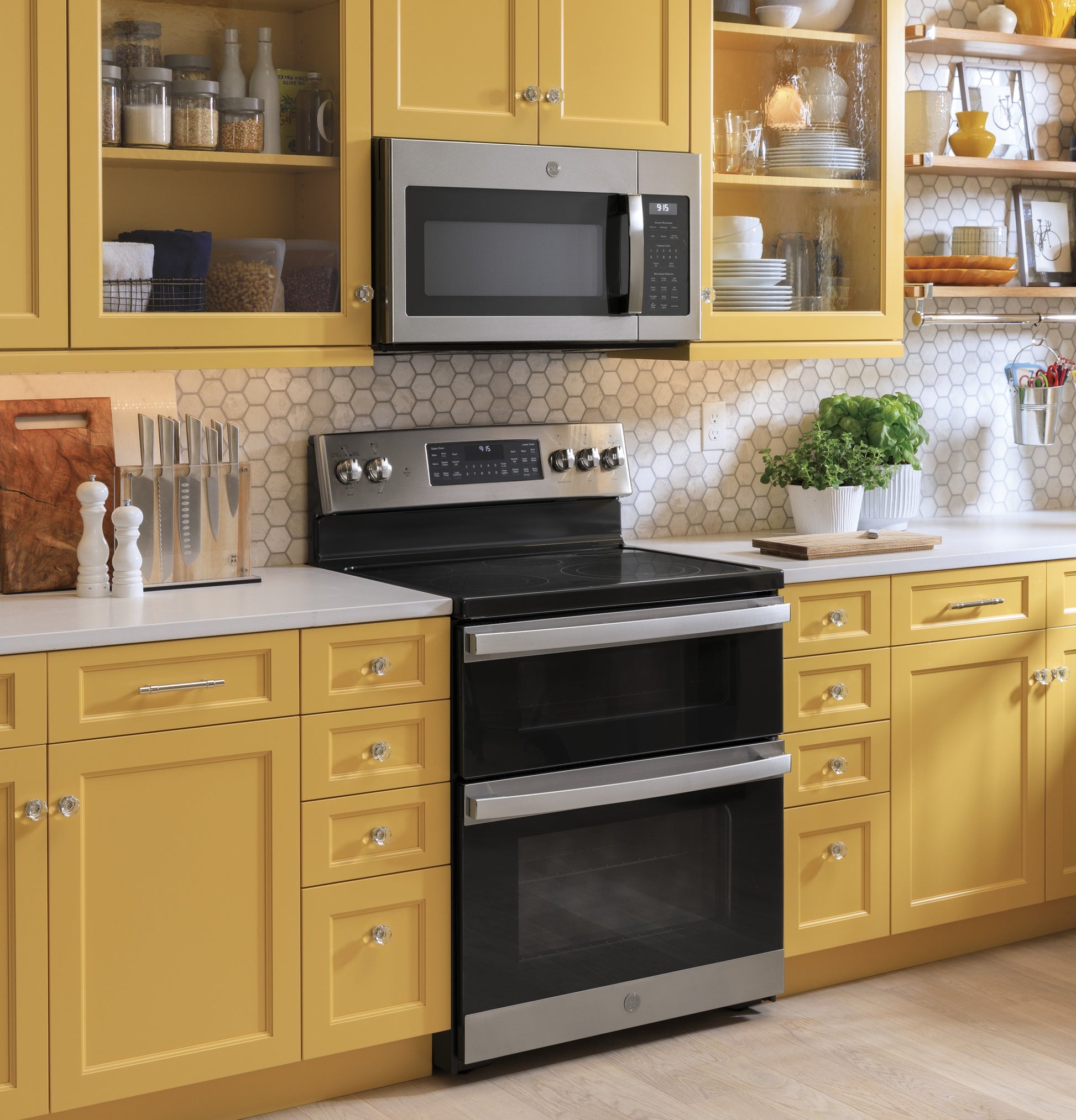 Stainless steel over-the-range microwave above black double oven in yellow kitchen with hex tile backsplash and plants