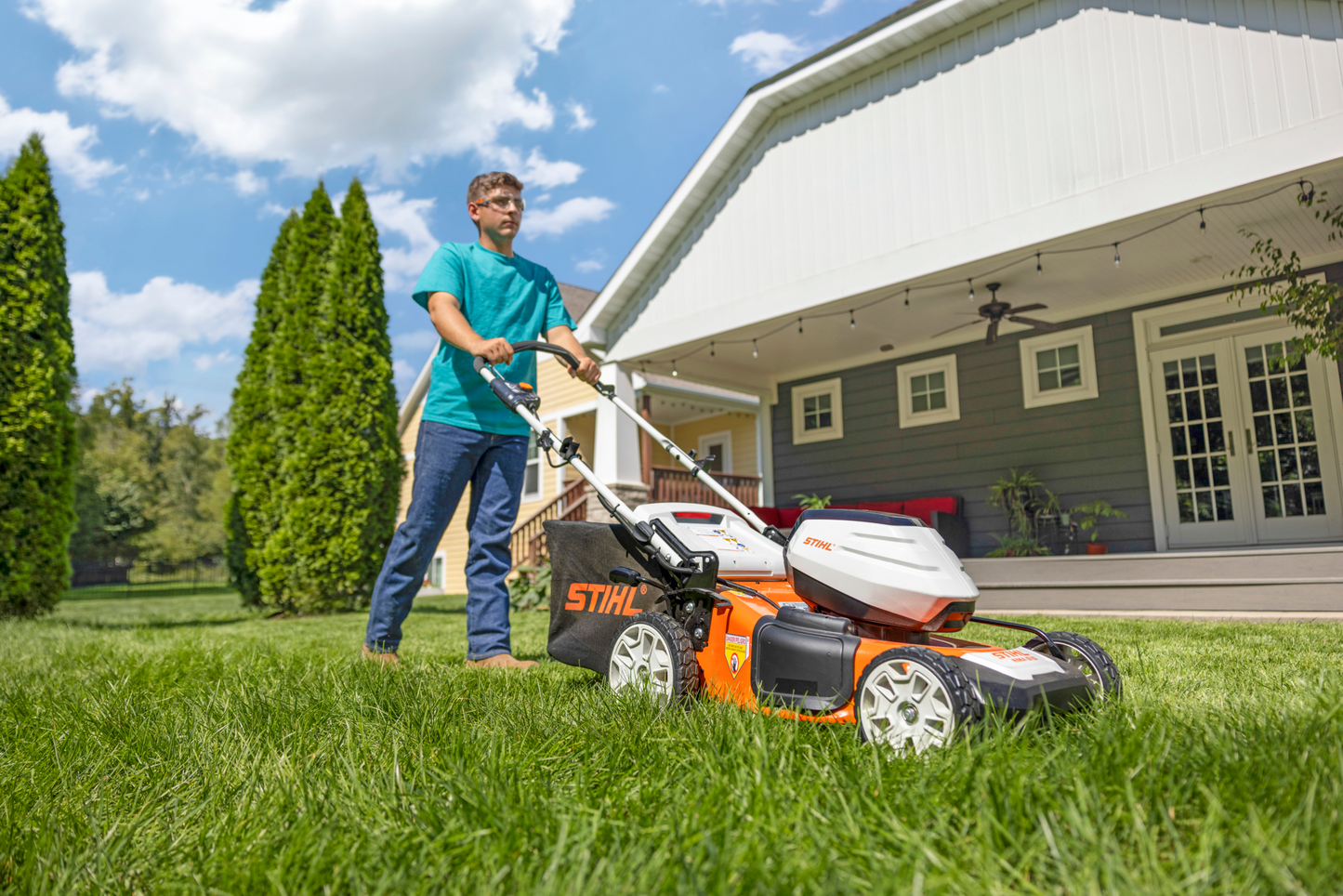 Man mowing lawn with orange and white STIHL battery-powered mower on green grass outside a modern house