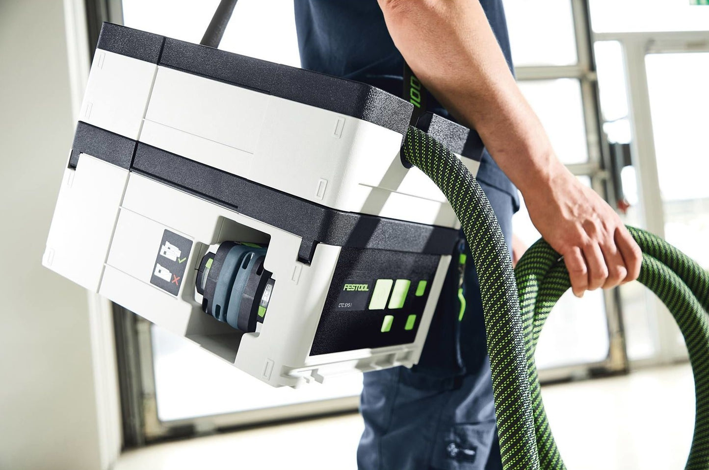 Person carrying a compact Festool cordless dust extractor with green hose, part of woodworking tools collection