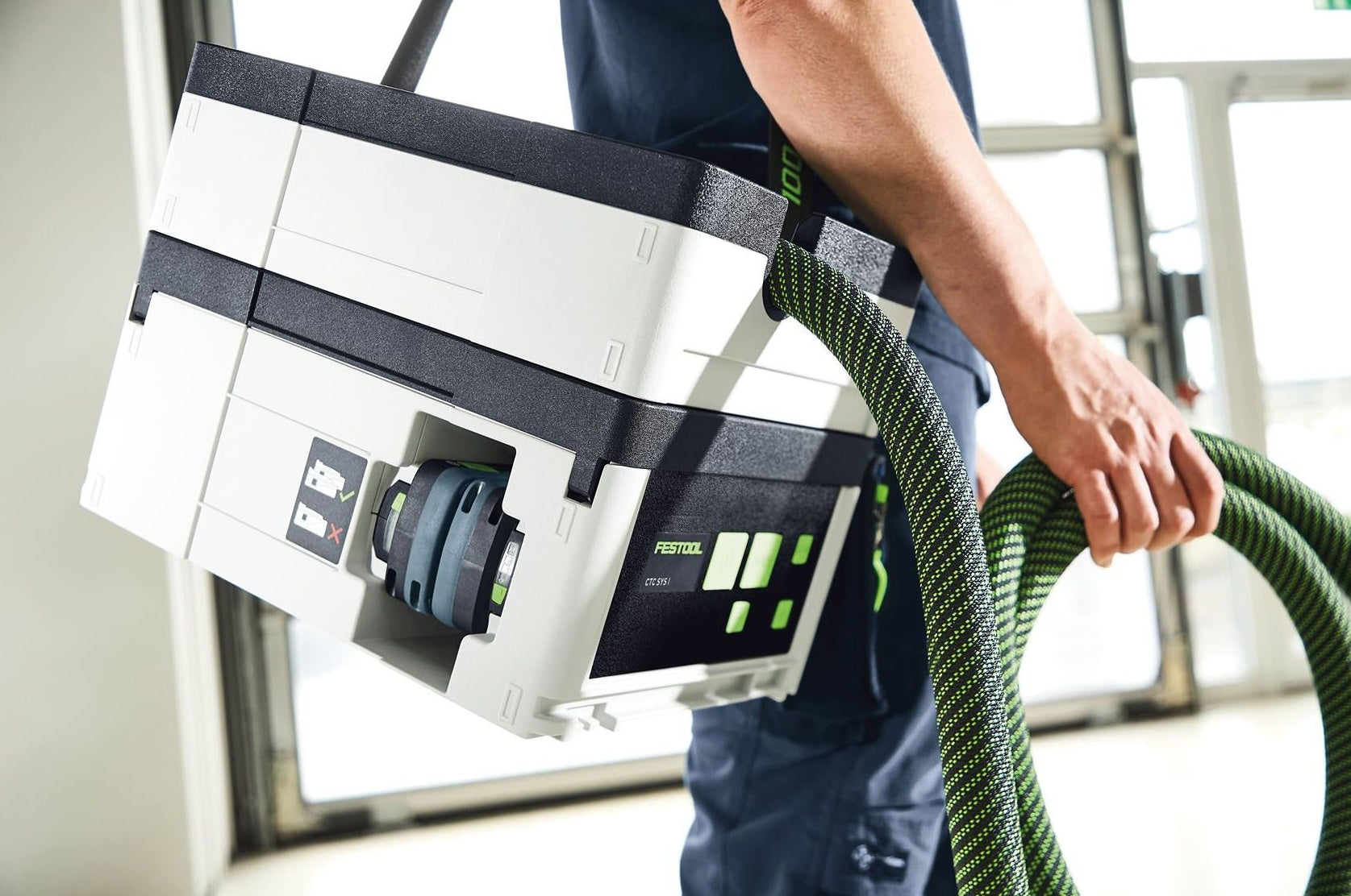 Person carrying a compact Festool cordless dust extractor with green hose, part of woodworking tools collection