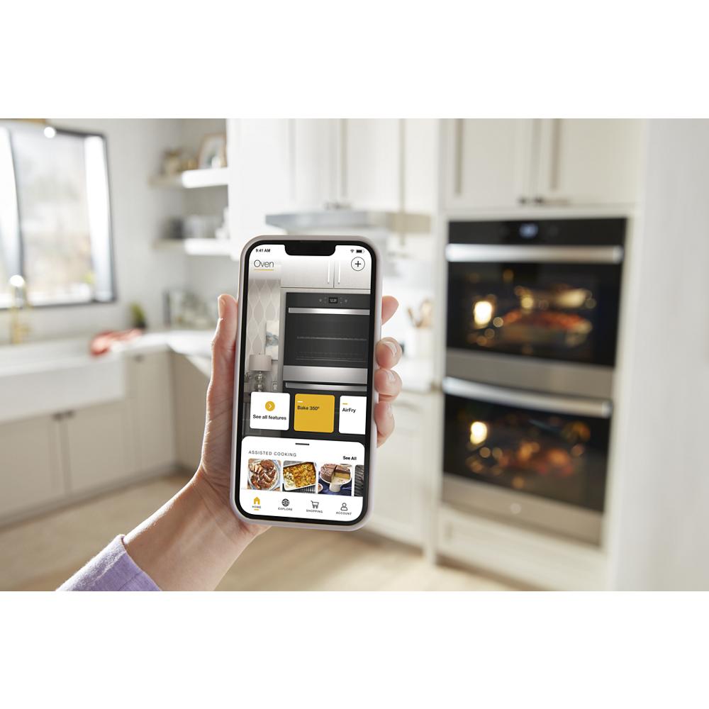 Person using smartphone app to control double wall oven remotely in modern kitchen with white cabinets