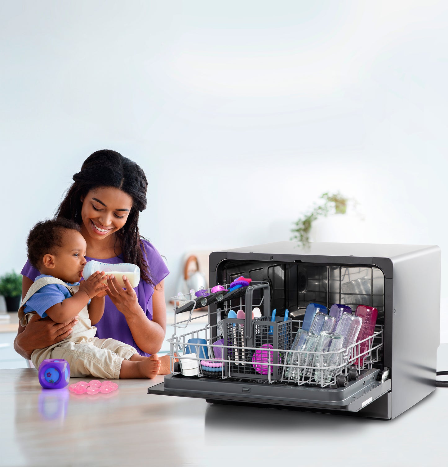 Compact dishwasher open with baby bottles and kitchenware next to smiling mother holding baby in bright kitchen setting