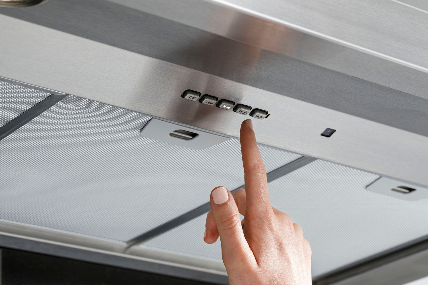 Close-up of hand pressing control button on stainless steel wall mount range hood in kitchen appliances collection