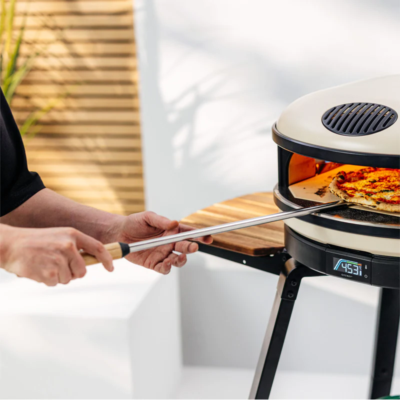 Person using a long-handled Gozney Turning Peel to remove a pizza from an outdoor pizza oven at 453 degrees