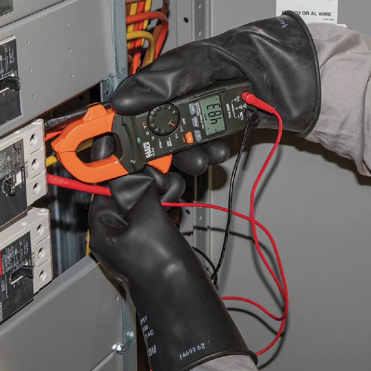 Person wearing protective gloves using a digital clamp meter for electrical testing in a panel box setting