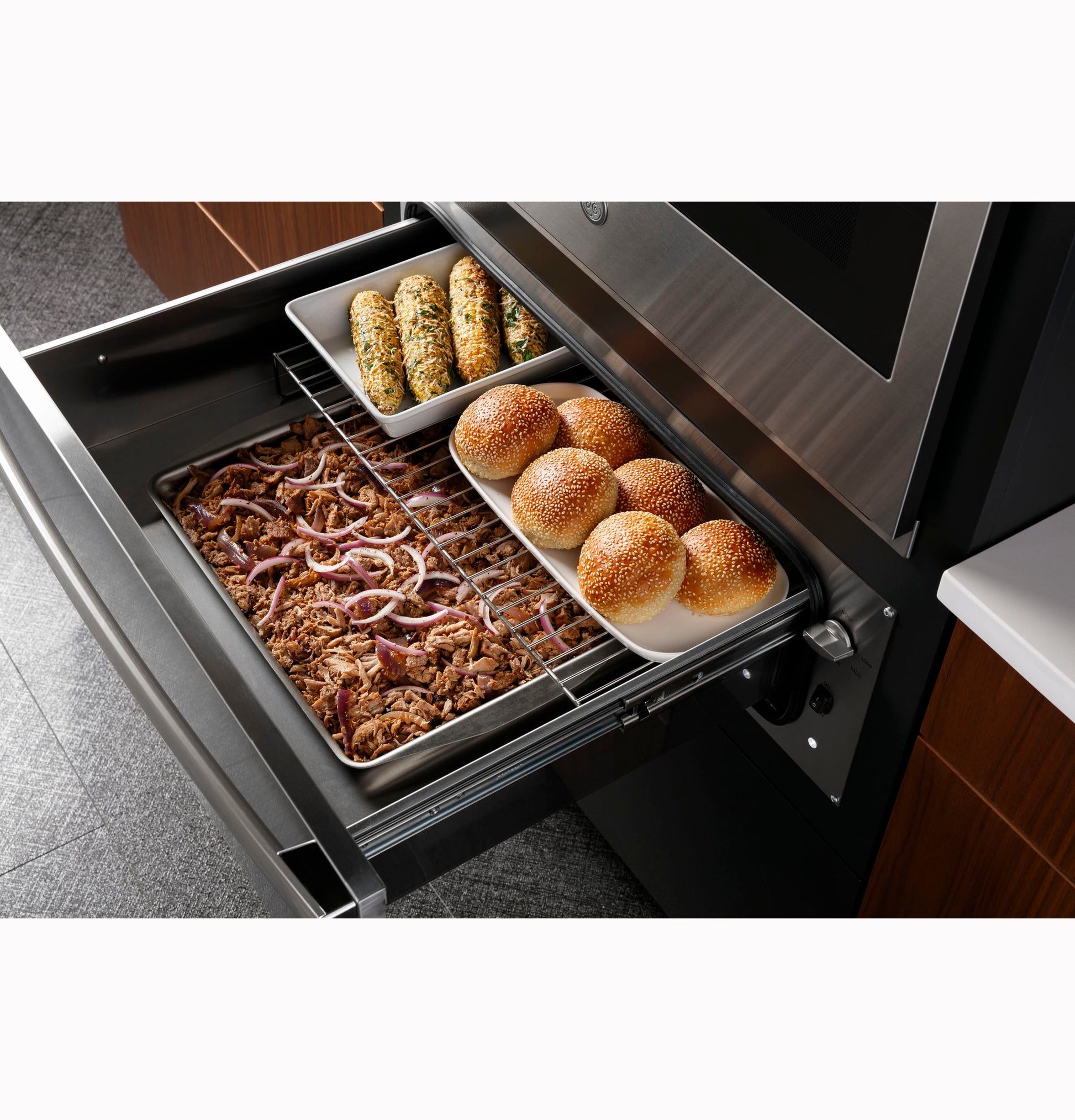 Open warming drawer with trays of pulled pork, bread rolls, and stuffed jalapeños in a modern kitchen setting