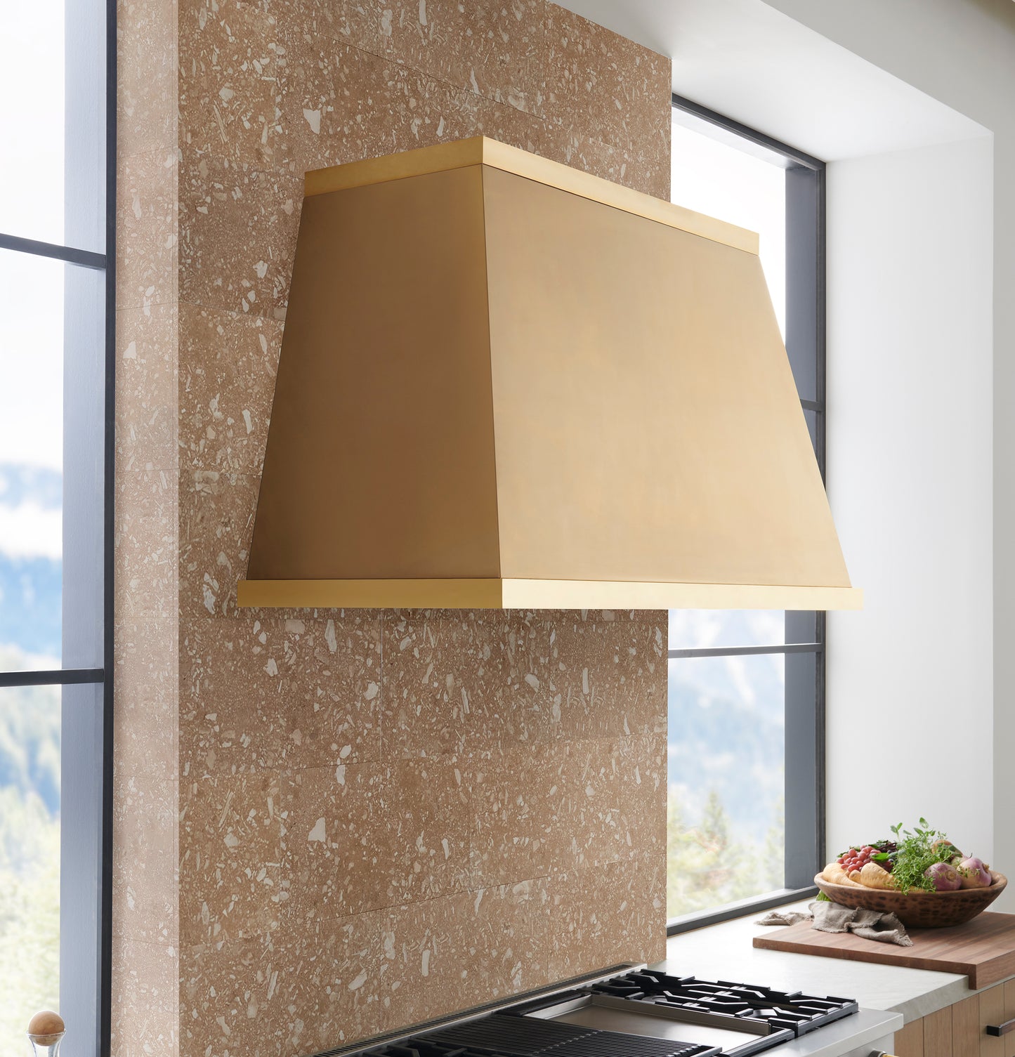 Minimalist brass kitchen hood mounted on textured stone wall with natural light from large windows in kitchenware collection