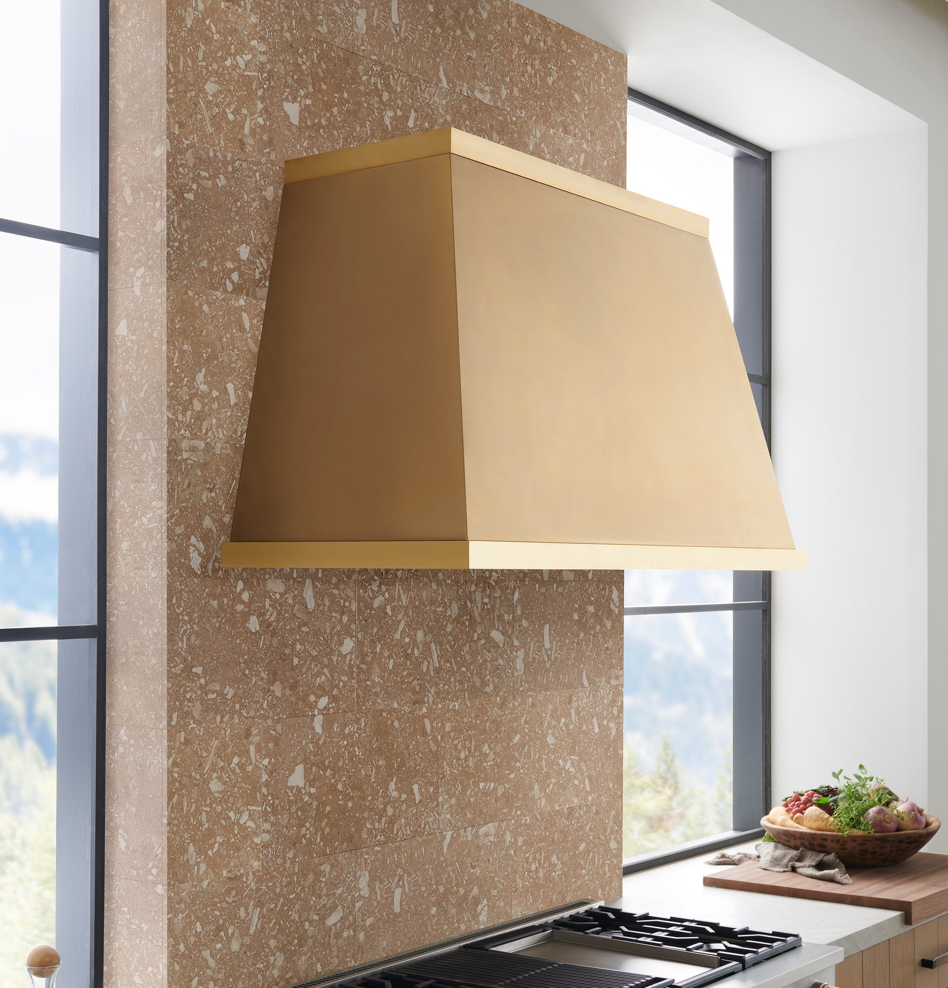 Minimalist brass kitchen hood mounted on textured stone wall with natural light from large windows in kitchenware collection