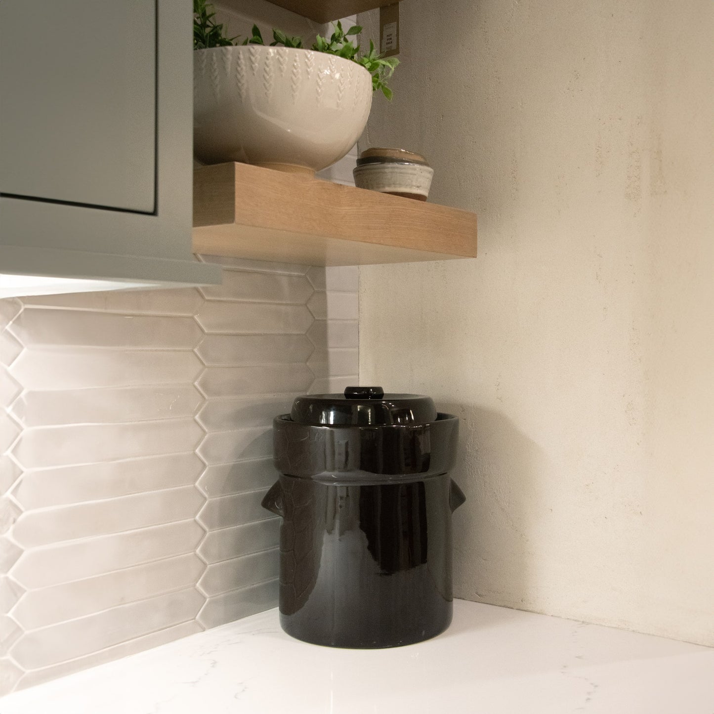 Glossy black ceramic crock with lid on white countertop in kitchen with tiled backsplash and wooden shelves above