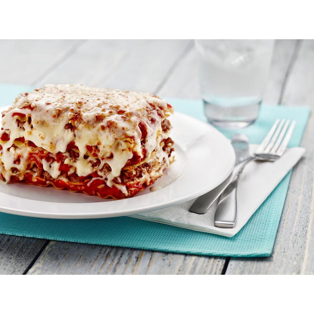 Close-up of cheesy lasagna slice on white plate with fork, knife, and glass of water on rustic wooden table
