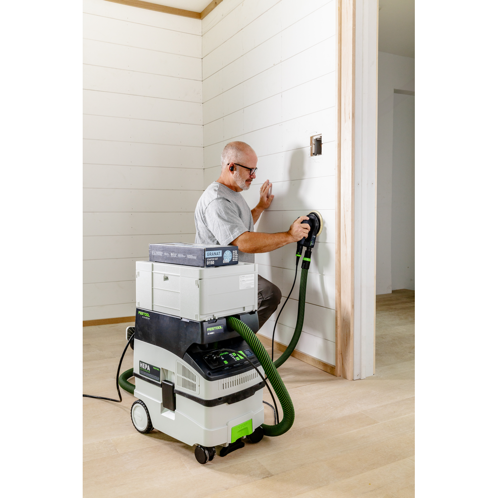 Man using a Festool sanding tool with a dust extractor on a white wood-paneled wall in a home improvement setting