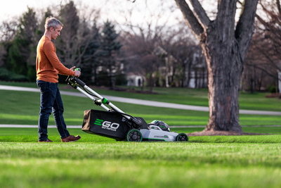 Person using a green and black cordless self-propelled mower on a residential lawn outdoors
