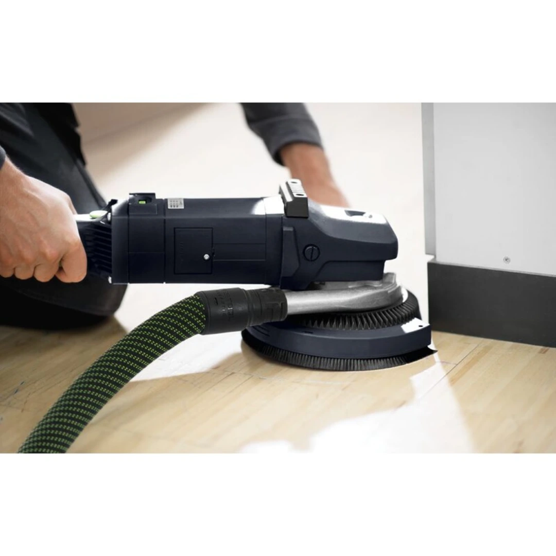 Handheld power sander with attached soft sanding pad smoothing a hardwood floor near a white baseboard