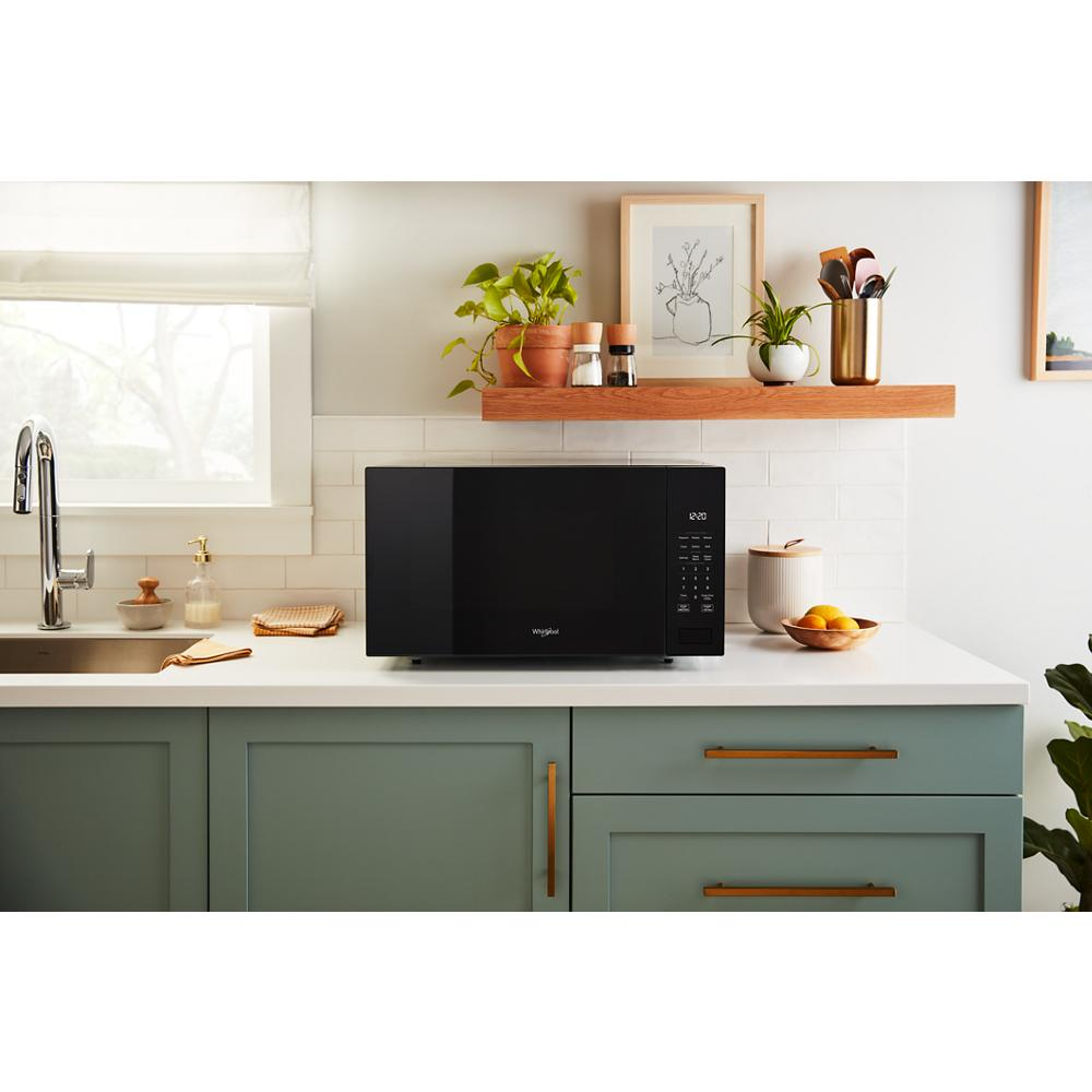 Black sensor cooking microwave with digital controls on white countertop in kitchen with green cabinets and wood shelf