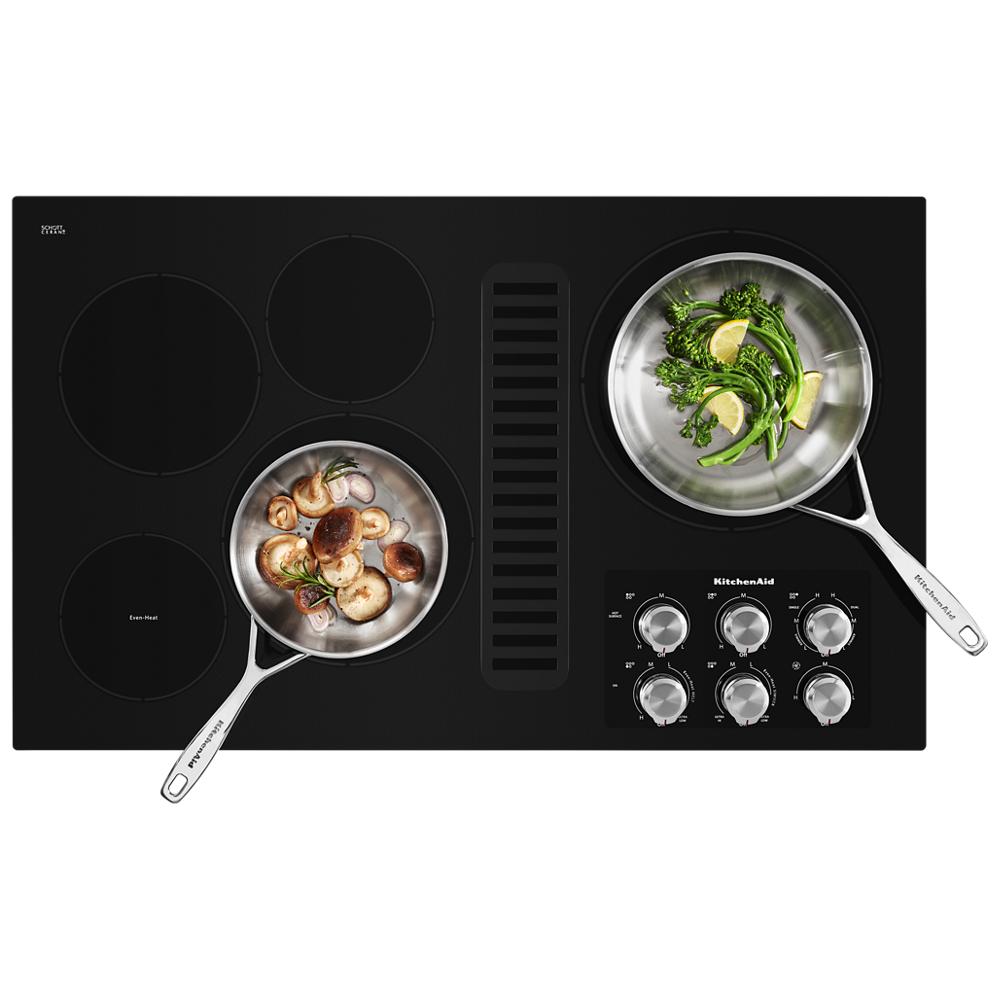 Black electric cooktop with five burners, downdraft vent, and stainless steel control knobs with pots cooking vegetables and mushrooms