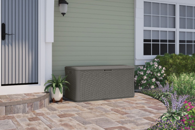 Brown resin wicker deck box with hinged lid placed on patio outside house near plants and window for home storage
