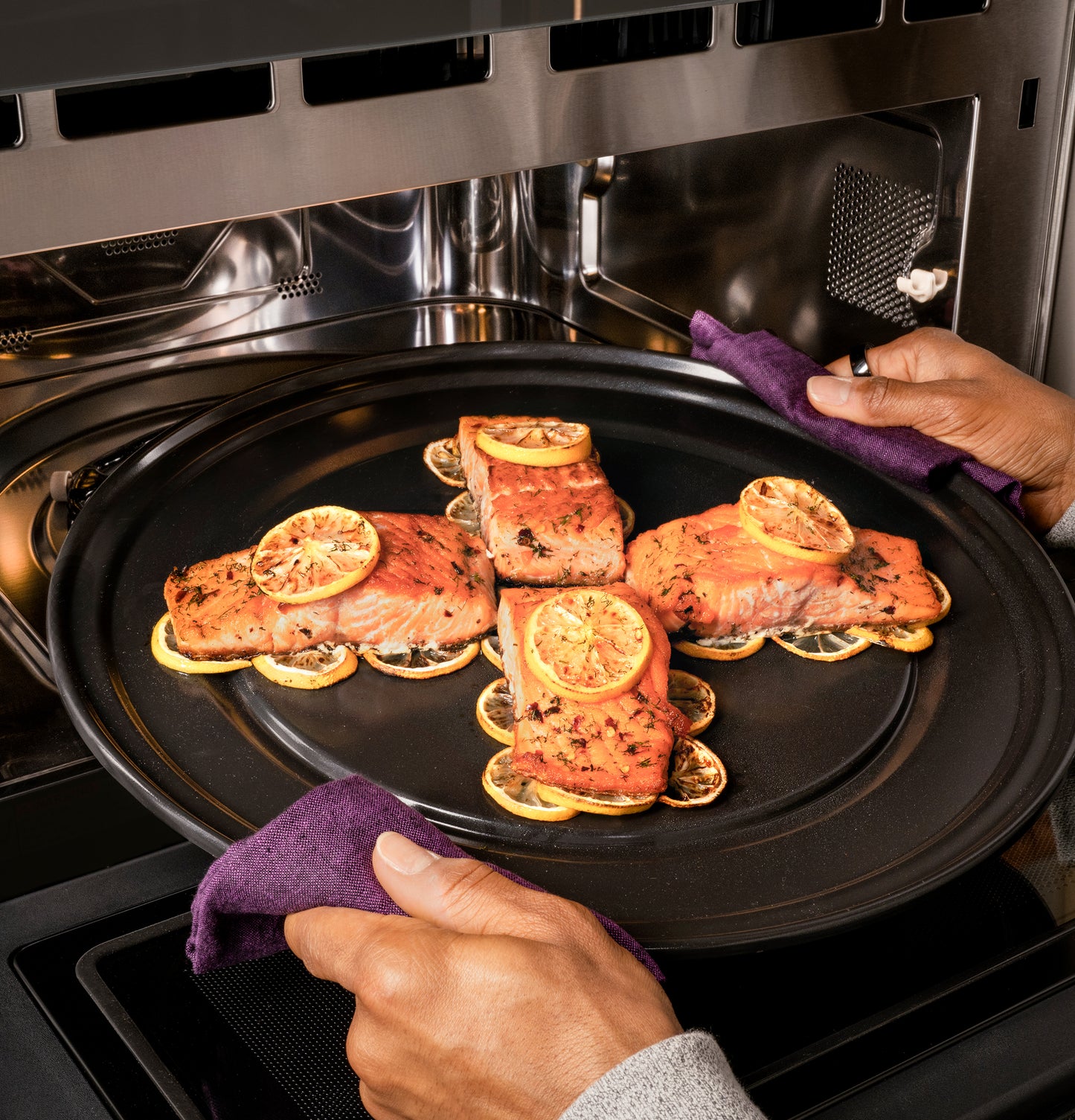 Person removing tray with cooked salmon topped with lemon slices from stainless steel wall oven in kitchen appliances setting
