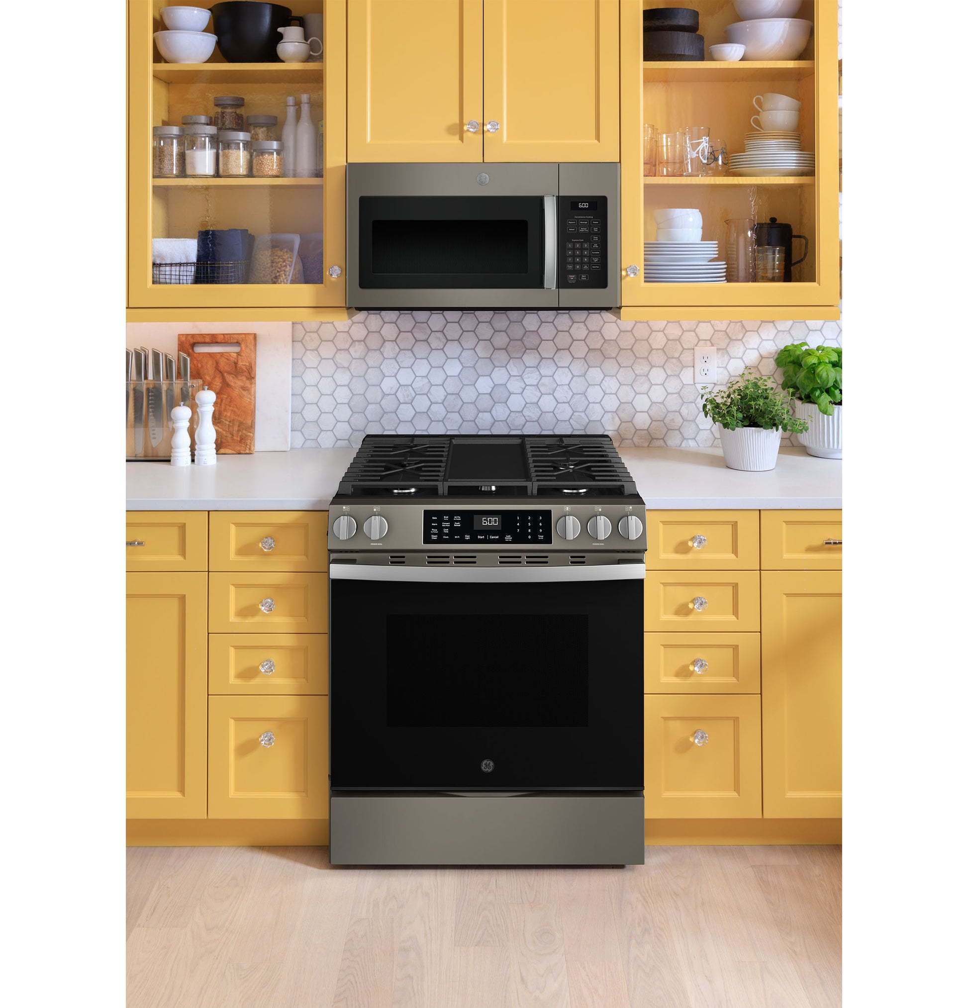 Stainless steel front-control gas range with convection oven and black microwave above in yellow kitchen cabinetry setting
