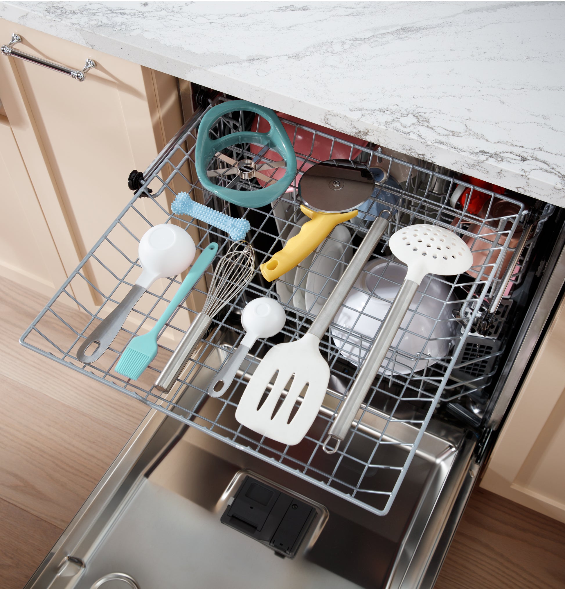 Top rack of stainless steel dishwasher holding kitchen utensils including spatula, whisk, and ladles inside modern kitchen cabinetry.