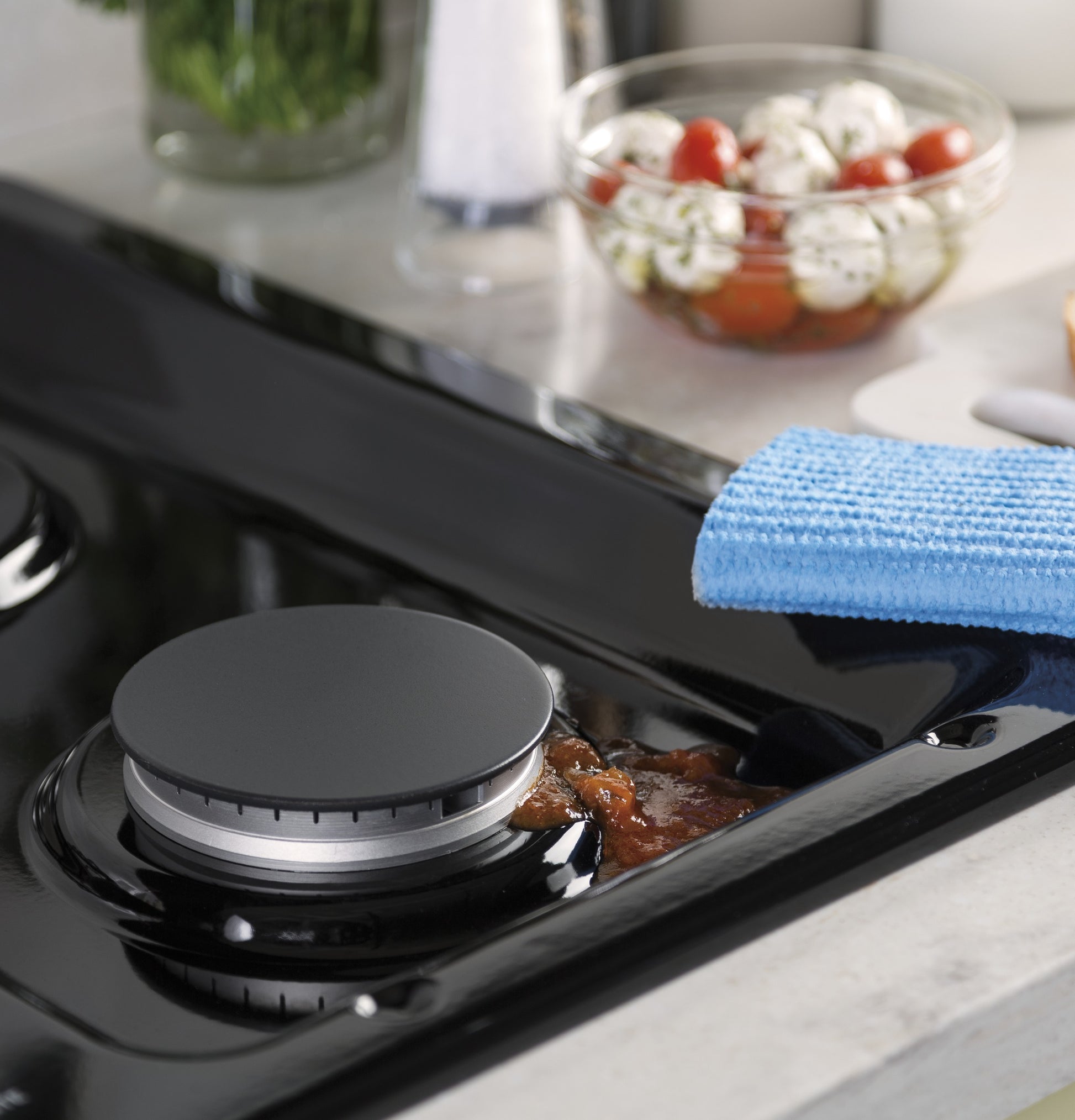Close-up of black gas cooktop burner with spilled sauce being wiped by blue cloth in modern kitchen setting