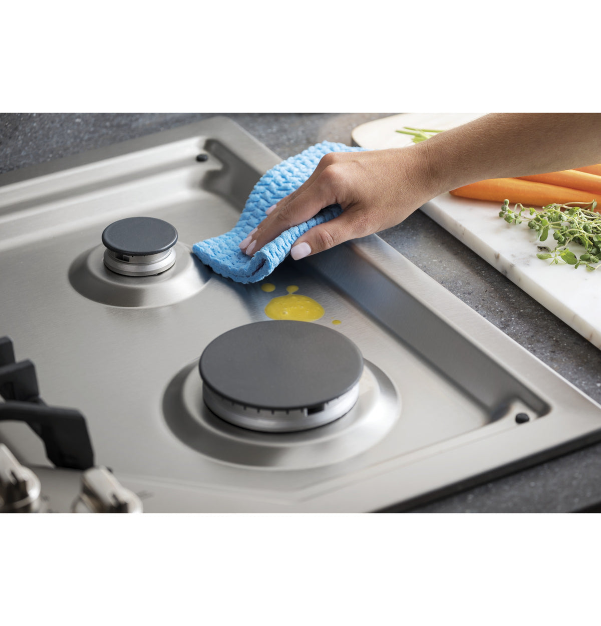 Close-up of a hand cleaning a stainless steel cooktop burner with a blue cloth in kitchen appliances collection