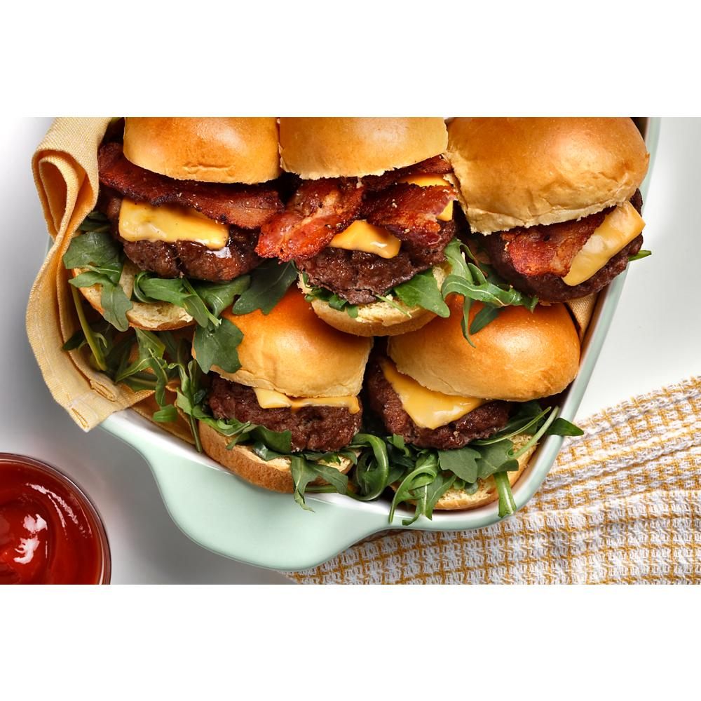 Close-up of small bacon cheeseburger sliders with lettuce on a white ceramic dish beside ketchup and kitchen towel
