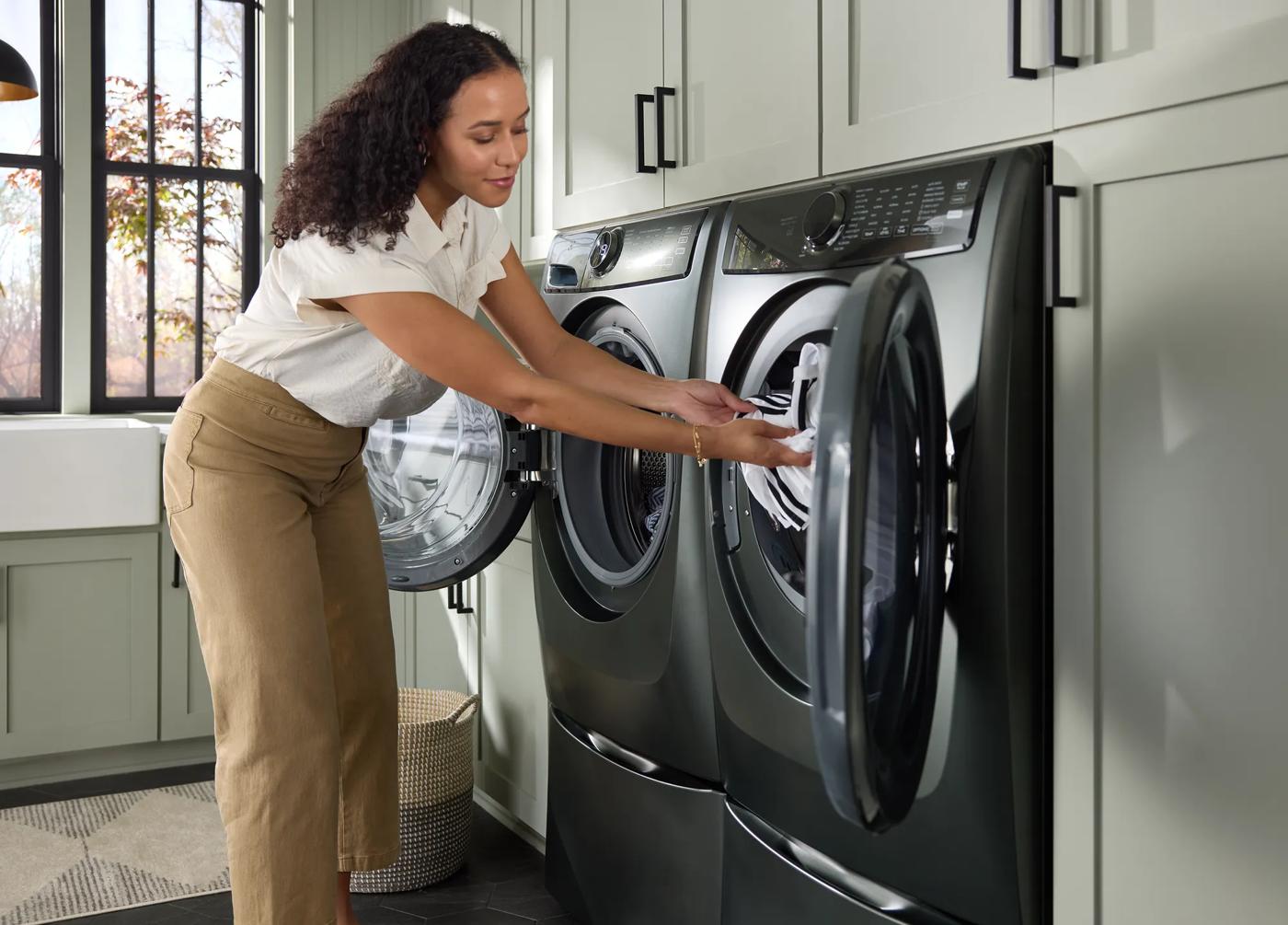 Woman loading clothes into a dark front load electric dryer in a modern laundry room with light gray cabinetry