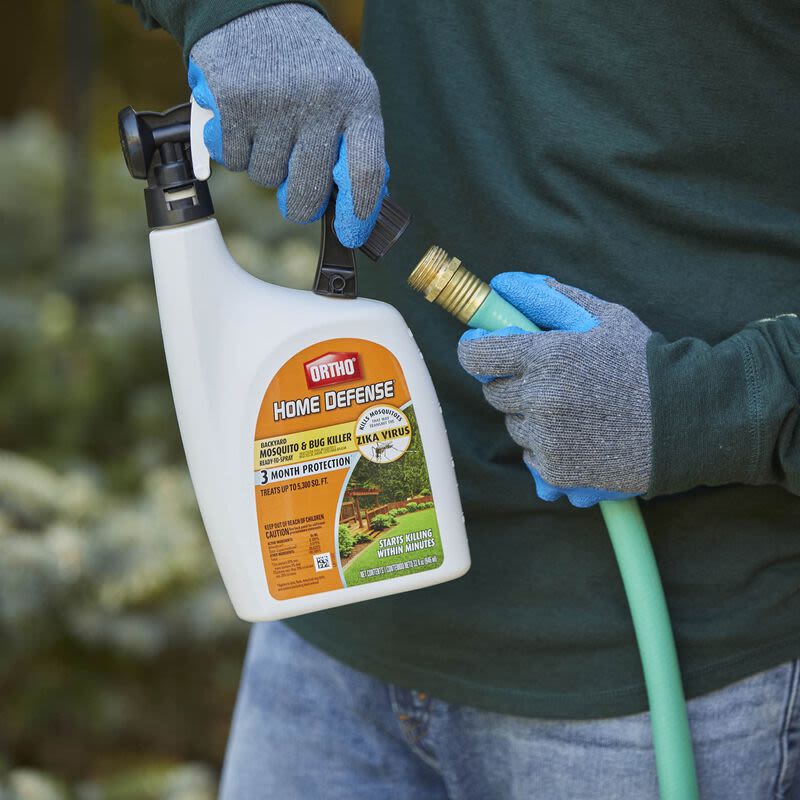 Person wearing gray gloves attaching Ortho Home Defense mosquito and bug killer spray to green garden hose outdoors