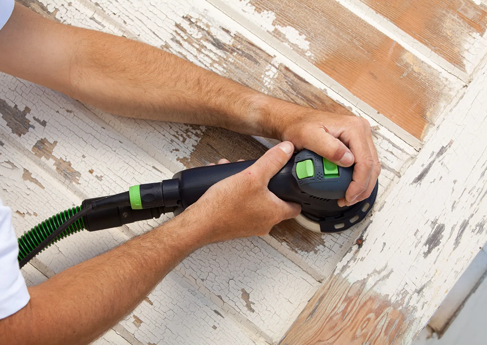 Hands using Festool dual mode sander on peeling paint wood siding for home improvement and woodworking tasks