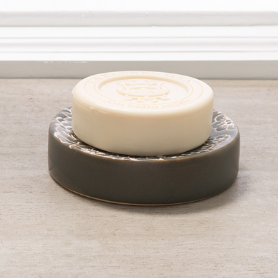 Dark ceramic soap dish with floral design holding a round cream-colored soap bar by a window sill