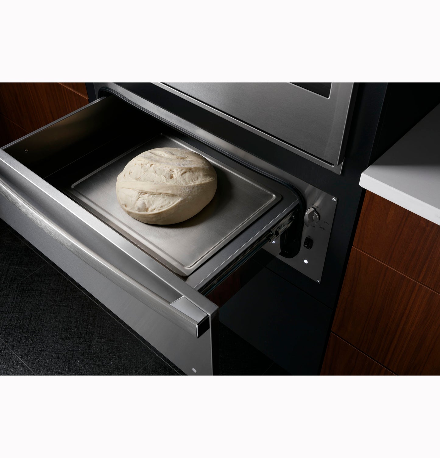 Open stainless steel warming drawer showing dough on a baking tray in modern kitchen setting