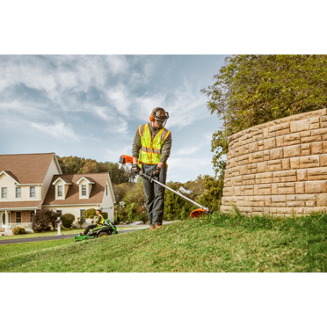 Worker in safety vest using a straight shaft trimmer to edge grass near a stone retaining wall on a sunny day