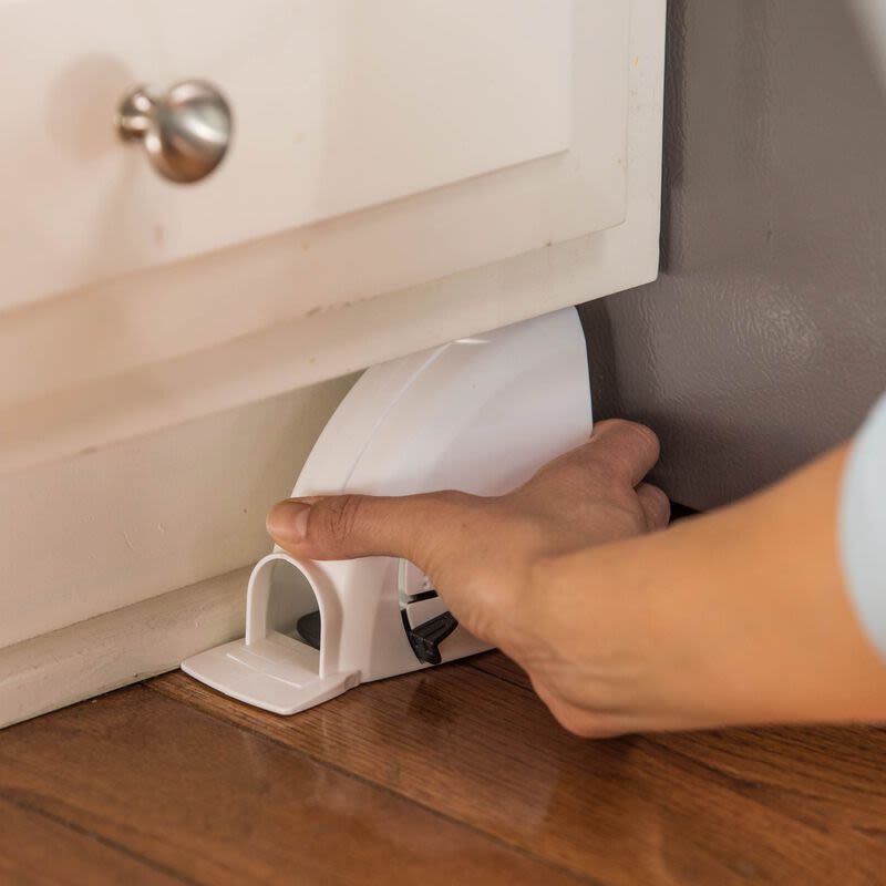 Hand setting a white plastic mouse trap on a hardwood floor next to a kitchen cabinet for pest control use