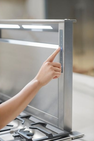Hand adjusting digital control panel on stainless steel downdraft ventilation unit above gas cooktop in kitchen setting