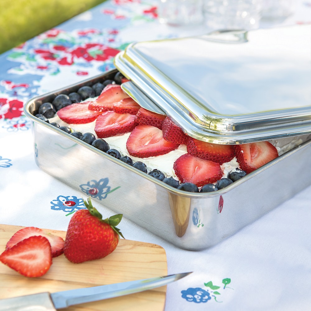 Stainless steel cake pan partially covered with lid, filled with dessert topped with strawberries and blueberries on floral cloth