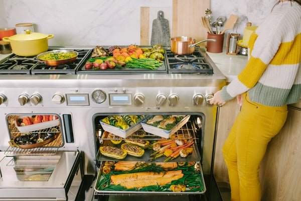 Person adjusting knob on dual fuel professional range with multiple dishes cooking in oven and on stovetop