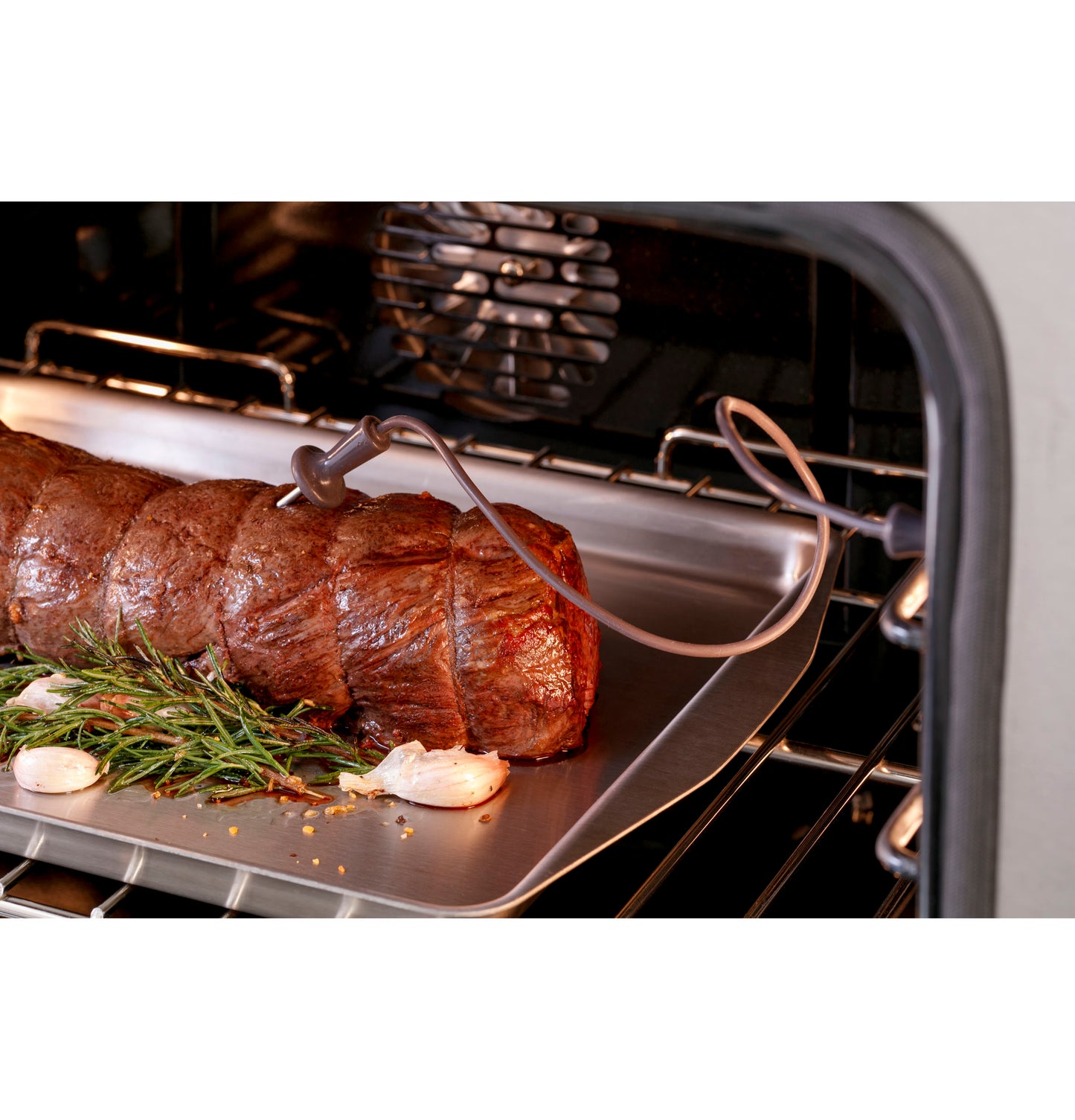 Beef roast cooking in oven with meat thermometer probe inserted alongside herbs and garlic cloves