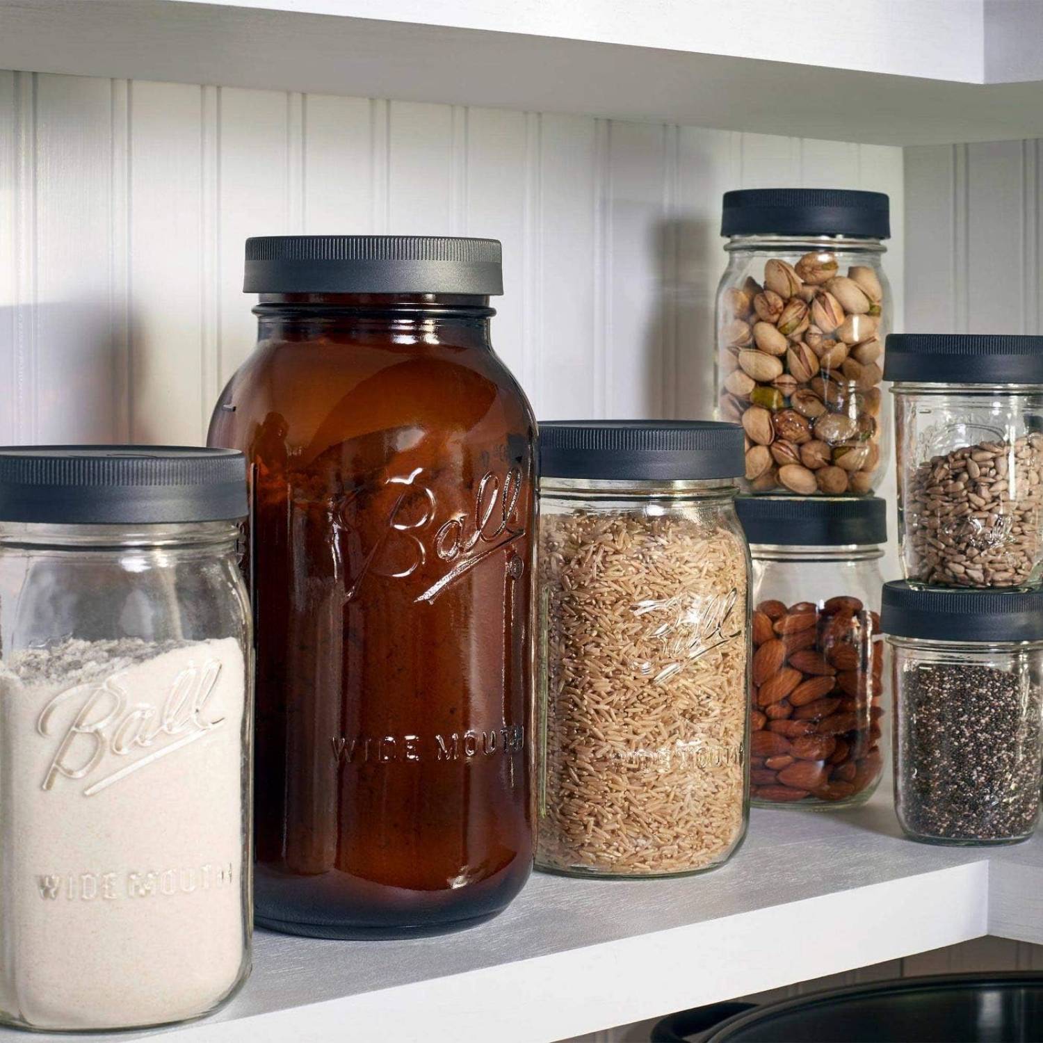 Assorted wide-mouth glass jars with black lids containing various grains, nuts, and seeds on a kitchenware shelf